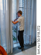 A contemplative teenage boy gazes out of a high-rise apartment window at the urban cityscape, his view partly obscured by sheer curtains. Стоковое фото, фотограф Данила Большаков / Фотобанк Лори