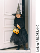 Happy laughing child girl in witch costume with a pumpkin basket for sweets in her hands at home near the door and is going to ask for candy while visiting friends and neighbors on Halloween night. Стоковое фото, фотограф Ирина Михайличенко / Фотобанк Лори