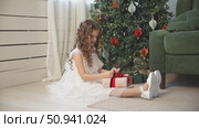 Cute happy little girl with curly hair, in an elegant white fluffy dress sits on floor under decorated Christmas tree at home, joyfully opening a gift box. Childhood and happy family holidays concept. Стоковое видео, видеограф Ирина Михайличенко / Фотобанк Лори