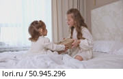 Two sisters wearing white dresses, sitting on a bed with white bedding, pulling big wrapped gift box, fighting, cannot share a present on the holiday morning at home. Стоковое видео, видеограф Ирина Михайличенко / Фотобанк Лори