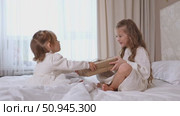 Two sisters wearing white dresses, sitting on a bed with white bedding, pulling big wrapped gift box, fighting, cannot share a present on the holiday morning at home. Стоковое видео, видеограф Ирина Михайличенко / Фотобанк Лори