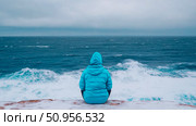 A lone figure in a vibrant blue jacket sits facing the vast, turbulent ocean under an overcast sky, contemplating the immense power of nature from a snowy cliff edge. Стоковое AI-изображение, AI-художник Данила Большаков / Фотобанк Лори