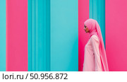 Graceful woman in a soft pink hijab and attire, standing against a vibrant turquoise and fuchsia abstract wall; a modern fashion concept. Стоковое AI-изображение, AI-художник Данила Большаков / Фотобанк Лори