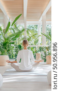 A serene woman meditating in a bright, sun-drenched wellness space surrounded by abundant tropical plants, for calm and inner peace. Стоковое AI-изображение, AI-художник Данила Большаков / Фотобанк Лори