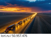 A convoy of massive semi-trucks travels closely together on a highway. The impressive scene unfolds under a dramatic dusk sky, highlighting the significance of road freight transportation. Стоковое AI-изображение, AI-художник Nart / Фотобанк Лори