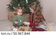 Two little girls, an older sister and a toddler, sit on the floor under the Christmas tree at home, playing with gifts and toys. Happy family and joy of winter holidays celebration concept. Footage. Стоковое видео, видеограф Ирина Михайличенко / Фотобанк Лори