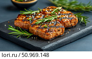 Three perfectly grilled and seasoned pork tenderloin medallions or steaks with distinct grill marks, garnished with fresh rosemary and white sesame seeds, elegantly arranged on a dark slate serving board against a muted background with blurred herbs and a bowl, showcasing a gourmet meal. Стоковое AI-изображение, AI-художник Данила Большаков / Фотобанк Лори