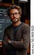 Stylish man with curly hair, modern glasses, and a full beard, wearing a brown knit cardigan with arms crossed, exuding confident and approachable masculinity in an indoor setting. Стоковое AI-изображение, AI-художник Данила Большаков / Фотобанк Лори