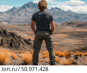 A man stands with his back to a majestic mountain landscape. He is wearing a dark T-shirt and hiking pants with pockets, ideal for trekking and outdoor activities. The dry vegetation in the foreground and the mountain range on the horizon emphasize the atmosphere of freedom, strength and travel. A perfect image for themes related to adventure, nature, tourism and personal freedom. Стоковое AI-изображение, AI-художник Анна Кривицкая / Фотобанк Лори