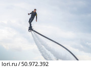 A man in a black athlete wetsuit hovers against a dramatic sky on a flyboard. A performer demonstrates impressive flyboarding skills, soaring above the clear blue water. Dressed in a sleek wetsuit, Стоковое фото, фотограф Владимир Разгуляев / Фотобанк Лори