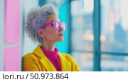 Confident woman with vibrant pink glasses and curly grey hair, dressed in bright yellow and fuchsia, looks contemplatively upwards in a bright, modern interior. Стоковое AI-видео, аниматор Данила Большаков / Фотобанк Лори