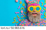 Joyful bearded man in a whimsical, sprinkle-covered hat and oversized candy glasses, smiling brightly against a vibrant blue background adorned with playful confectionery shapes. Стоковое AI-изображение, AI-художник Данила Большаков / Фотобанк Лори