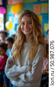 Smiling young woman with wavy blonde hair and crossed arms, radiating warmth and approachability in vibrant, blurred classroom filled with children in background. Стоковое AI-изображение, AI-художник Данила Большаков / Фотобанк Лори