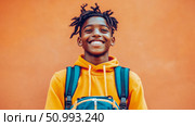 Portrait of a smiling adolescent boy with dreadlocks wearing a yellow hoodie and backpack against an orange background. Стоковое AI-изображение, AI-художник Данила Большаков / Фотобанк Лори