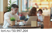 Young man is sitting at a table in a cafe and working at a laptop. Guy designer works remotely in a coffee shop. Стоковое видео, видеограф Яков Филимонов / Фотобанк Лори