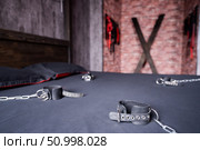 Torture of Andreev cross on the bedroom wall. VDSM equipment and sex toys. Leather handcuffs with a chain on a gray sheet. The lashes hang in the room for perversions. Стоковое фото, фотограф Михаил Решетников / Фотобанк Лори