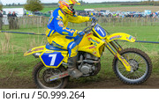 Motorcycle racer in bright yellow gear navigates a muddy track, showcasing skill and speed during an off-road competition with spectators in the background. Стоковое AI-изображение, AI-художник Ирина Ручушкина / Фотобанк Лори