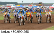 Group of motocross riders in colorful gear racing on dirt track, showcasing speed and competition in an outdoor event with spectators and tents in the background. Стоковое AI-изображение, AI-художник Ирина Ручушкина / Фотобанк Лори