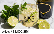 A refreshing Hugo Spritz cocktail in a glass with ice, elderflower, mint leaves, and lime slices. A bottle of prosecco is visible in the background. Стоковое AI-изображение, AI-художник Журавлева Кристина / Фотобанк Лори