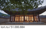 Soft illumination glowing through wooden lattice doors of traditional Korean Hanok, dark tiled roof framed by stone wall and verdant greenery during twilight hours. Стоковое AI-видео, аниматор Данил Руденко / Фотобанк Лори