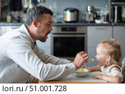 A caring man with a beard feeds his little daughter. A little girl sits at a children's dining table and dines with her dad. Happy fatherhood. Стоковое фото, фотограф Михаил Решетников / Фотобанк Лори