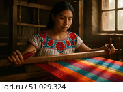 Focused young Mexican artisan in a white blouse with red and blue embroidery weaving rainbow-colored fabric on a wooden loom. Стоковое AI-изображение, AI-художник Башкирова Анна / Фотобанк Лори