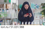 Young Muslim woman in traditional islamic navy blue dress and niqab covering face except eyes, shopping for medications or care products in drugstore, using phone to check list of needed items. Стоковое видео, видеограф Яков Филимонов / Фотобанк Лори