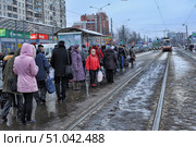 Winter cityscape in St. Petersburg with a tram stop. In the foreground there is a queue of people in warm winter clothes waiting for the tram to arrive. Many pedestrians are visible, some with bags and packages, ready for the ride. In the background a red and white tram moves along snowy and wet rails. On both sides of the street there are shops and trade pavilions, among which the signs "Megafon" and "Denga" are noticeable. Soviet-built houses and modern buildings create a contrast of urban architecture. The atmosphere of a gray winter day with wet asphalt and remnants of snow on the roadsides conveys the everyday life of a metropolis. (2013 год). Редакционное фото, фотограф Анна Кривицкая / Фотобанк Лори