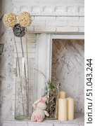 Cozy winter interior with decorative elements in light colors. In the foreground is a fragment of a snow-white fireplace with brick trim and a carved cornice. On the left, in a tall glass vase, there are decorative spherical flowers on long stems in cream and silver colors, at the base of which are twisted silver branches and a miniature pendant in the form of a Christmas tree. Nearby on the shelf sits a soft toy - a white plush hare with pink paws and ears. On the right, in the middle glass vase, there is a composition of spruce branches, driftwood and decorative shiny elements. Nearby are three cream cylindrical candles. The background is made of gray and white textured decorative plaster, creating an atmosphere of winter comfort and lightness. Стоковое фото, фотограф Анна Кривицкая / Фотобанк Лори