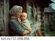 Деревенская жизнь: бабушка у дома держит на руках внука-малыша. Village life: a grandmother holding a baby grandson in her arms. Стоковое AI-изображение, AI-художник Смирнова Светлана / Фотобанк Лори