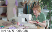 Mature female seamstress is sitting near sewing machine and create new dress in the workshop. Стоковое видео, видеограф Яков Филимонов / Фотобанк Лори