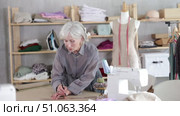 Attentive old tailoress recording dress design on large paper in sewing studio. Стоковое видео, видеограф Яков Филимонов / Фотобанк Лори