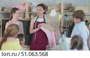Friendly female sewing teacher using chalkboard pattern and mannequin to explain garment construction to group of school-age students seated around table in classroom. Стоковое видео, видеограф Яков Филимонов / Фотобанк Лори