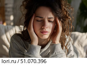 Красивая молодая женщина с головной болью сидит на диване, обхватив голову обеими руками и слегка щурясь от боли. Крупный план. Стоковое AI-изображение, AI-художник Максим Коломыченко / Фотобанк Лори