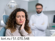 Красивая молодая женщина сидит в современном стоматологическом кресле, приложив руку к щеке от зубной боли, с рядом стоящим мужчиной-стоматологом в белом халате. Стоковое AI-изображение, AI-художник Максим Коломыченко / Фотобанк Лори