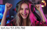 Excited female gamer celebrating victory with headphones in neon-lit room, expressing joy and energy in a dynamic gaming or streaming environment. Стоковое AI-изображение, AI-художник Катерина Бондарец / Фотобанк Лори