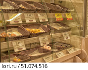 A bakery or supermarket display case with baked goods arranged in wicker baskets on multi-level shelves behind glass. Each basket contains different types of buns, donuts, croissants or pies, some of which have already been sold, leaving half-empty trays. In front of each basket are price tags indicating the cost and name of the products, some are highlighted with yellow and red marks indicating a promotion or discount. Warm light gives the image a cozy and appetizing atmosphere, emphasizing the golden color of the baked goods. (2015 год). Редакционное фото, фотограф Анна Кривицкая / Фотобанк Лори