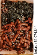 A photograph of souvenir keychains made in the form of miniature figures. In the upper part of the frame are keychains made of dark material depicting seated elephants, and in the lower part are keychains in the form of anatomically stylized figures in a brown wood-like design. The keychains are attached to metal key rings, lie in a box, forming a dense array, motley in colors and shapes. The products have a characteristic handmade look, with small surface irregularities, which gives them an authentic and handicraft style. Стоковое фото, фотограф Анна Кривицкая / Фотобанк Лори