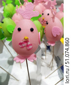 A bright decorative figurine in the form of a chicken, made in pink tones. The base is a soft pink plastic egg, decorated with eyes made of black beads, a small yellow beak and a pink fabric checkered bow. A comb made of pink felt is attached to the top, and wings and legs made of the same material are at the bottom and on the side. The figurine is mounted on a wooden skewer, which allows it to be used as a festive decoration or an element of the composition. In the background, other similar figurines in pink and green colors are visible, slightly blurred, which creates the effect of depth. Стоковое фото, фотограф Анна Кривицкая / Фотобанк Лори