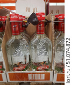 Bottles of Agavita Blanco tequila neatly arranged in boxes on a store shelf. Clear glass bottles with a bright red cap and a protective anti-theft device. The label depicts agave, a symbol of the drink's main ingredient, as well as inscriptions in Spanish indicating its origin in Mexico. At the bottom of the label is the word Tequila on a red background. The crystal clear contents are visible through the clear glass. In the background are repeating rows of bottles, creating a rhythmic composition. (2019 год). Редакционное фото, фотограф Анна Кривицкая / Фотобанк Лори