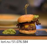 A delicious burger served on a black rectangular serving board. The burger includes a toasted sesame bun, a juicy beef patty, melted cheddar cheese, a fresh tomato slice, and green and purple lettuce leaves. The burger is held together with a wooden skewer for easy serving. A portion of avocado puree or guacamole sauce is neatly placed on the board next to it. The background is softly blurred, which emphasizes the sharpness and appetizing nature of the dish. The atmosphere of the photo conveys the coziness of restaurant serving and gastronomic pleasure. Стоковое фото, фотограф Анна Кривицкая / Фотобанк Лори