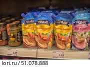 A shop window with a range of sauces in soft packaging from the Chumak brand. In the foreground are "French sauce with mustard" in yellow and blue packages and "Russian sauce with horseradish" in pink and blue packaging. The packages depict appetizing dishes - sausages with a side dish and meat with sauce. On the left in the frame, glass jars with other sauces and seasonings are visible. The price tags on the shelf indicate the cost of the products in euros and the price per kilogram. (2015 год). Редакционное фото, фотограф Анна Кривицкая / Фотобанк Лори