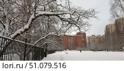 A winter cityscape with a snow-covered park and residential buildings in the background. The branches of the trees in the foreground are thickly covered with snow, creating an openwork pattern against the gray winter sky. On the left is a metal black fence, receding into the distance. In the center of the frame, a wide snow-covered area with benches and several passersby in warm winter clothes is visible. In the background, multi-story residential buildings made of red and beige brick rise. The atmosphere of the frame conveys the silence, coolness and tranquility of a winter day in the city. Стоковое фото, фотограф Анна Кривицкая / Фотобанк Лори