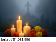 Candles and marigold flowers arranged in a cemetery. A cross is visible in the background, shrouded in fog. The scene represents Día de los Muertos traditions. Стоковое AI-изображение, AI-художник Nart / Фотобанк Лори