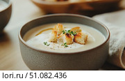 A bowl of creamy soup topped with croutons and herbs. The soup is served in a textured gray bowl on a wooden table with additional bowls in the background. Стоковое AI-видео, аниматор Nart / Фотобанк Лори