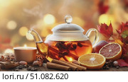 A glass teapot filled with steaming herbal tea sits on a wooden table. Slices of orange, cinnamon sticks, and autumn leaves surround the teapot, creating a warm atmosphere. Стоковое AI-видео, аниматор Nart / Фотобанк Лори