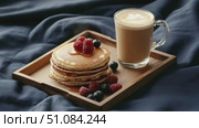 A wooden tray holds a stack of fluffy pancakes topped with raspberries and blueberries, alongside a glass of latte on a dark blue fabric background. Стоковое AI-видео, аниматор Nart / Фотобанк Лори