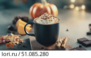 A black mug filled with pumpkin spice latte topped with whipped cream and caramel sits on a wooden board. Nearby are pumpkin slices, chocolate pieces, and autumn leaves. Стоковое AI-видео, аниматор Nart / Фотобанк Лори