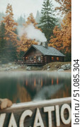 A cozy log cabin by a lake surrounded by autumn trees. A wooden sign reads 'VACATION'. Smoke rises from the chimney, creating a warm atmosphere. Стоковое AI-видео, аниматор Nart / Фотобанк Лори