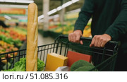 A person pushes a shopping cart filled with groceries in a supermarket. Items include a baguette, lettuce, cheese, and avocado. Brightly colored fruits are in the background. Стоковое AI-видео, аниматор Nart / Фотобанк Лори